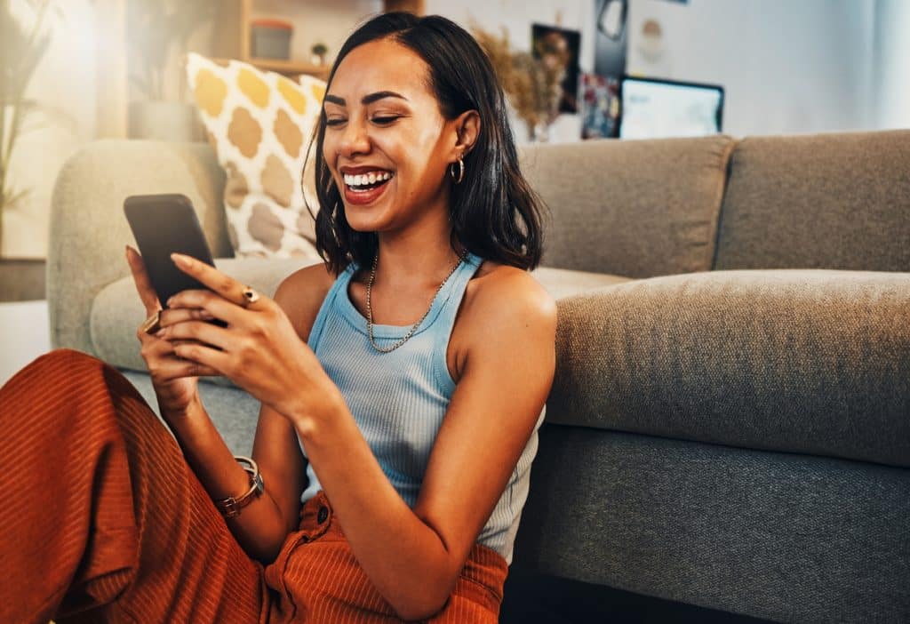 Tailored Ink | Copywriting and marketing agency | Beautiful mixed race woman browsing internet on cellphone in home living room. Happy hispanic sitting alone on floor in lounge and using technology to network. Laughing while scrolling on social media